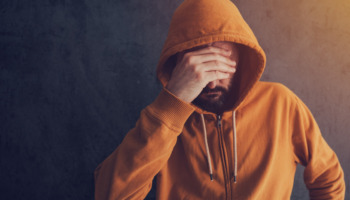 A man in an orange hoodie clutches his face, looking exhausted