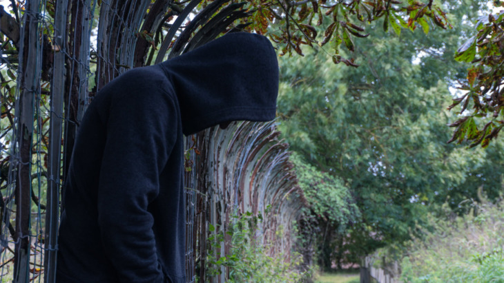 A depressed person in a hoodie leans against a fence