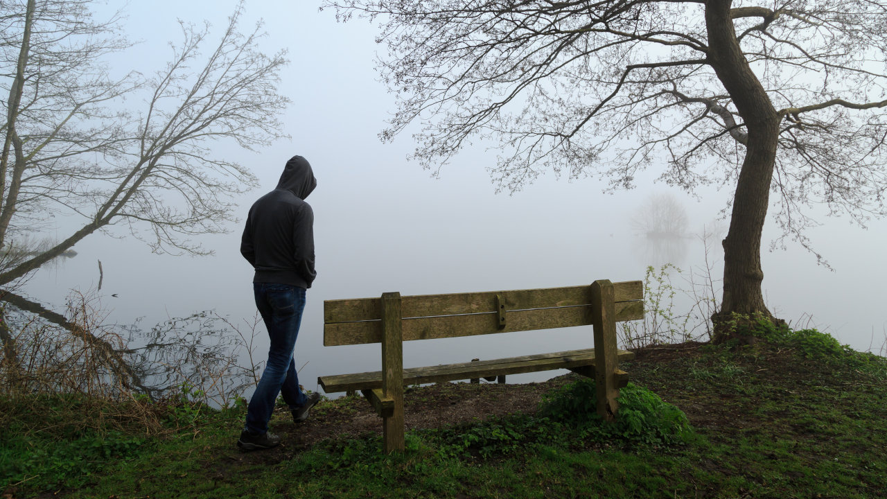 A depressed looking person in a hoodie approaches a bench on a gloomy day