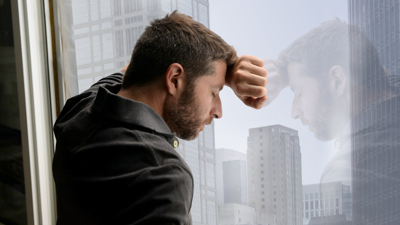 A depressed looking man rests against a window, looking out at the city