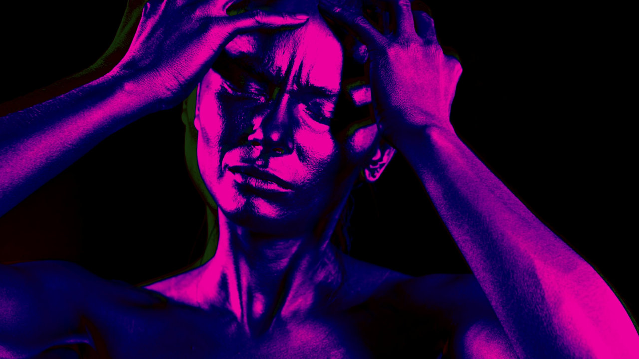 A woman bathed in purple lighting grips her forehead, possibly hallucinating.