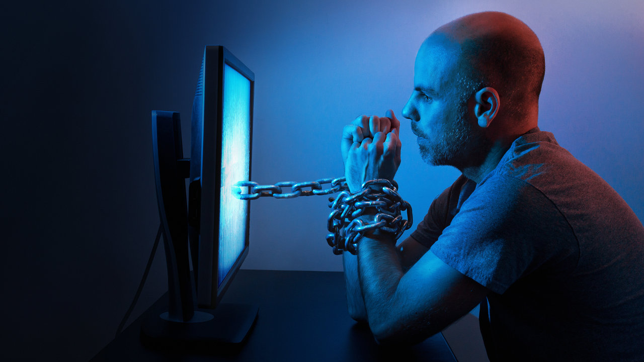 A man with his hands chained together, attached to a  computer screen, signifying computer or sex addiction.
