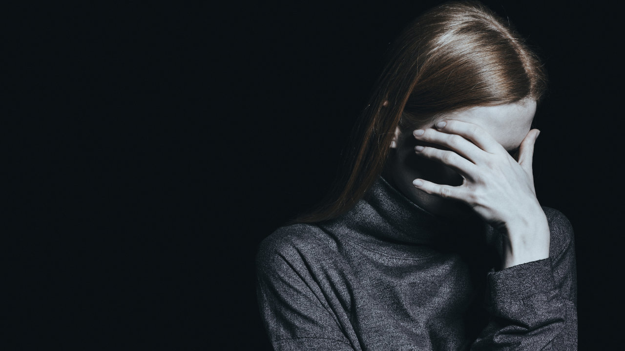 A woman covers her face, standing against a black background