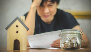 A stressed-looking man grips his forehead while looking at a jar of coins and a model house, suggesting difficulty paying a mortgage