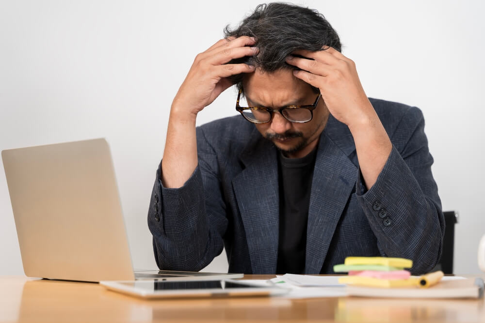 A businessman clutches his head while sitting at a laptop. Learn more about xanax addiction here.