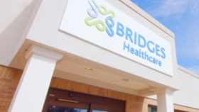 Exterior view of Bridges Healthcare facility entrance with illuminated logo sign, white facade, brick detailing, and clear blue sky