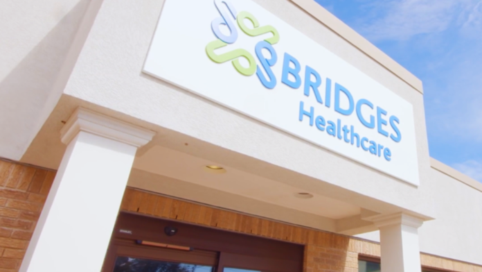 Exterior view of Bridges Healthcare facility entrance with illuminated logo sign, white facade, brick detailing, and clear blue sky