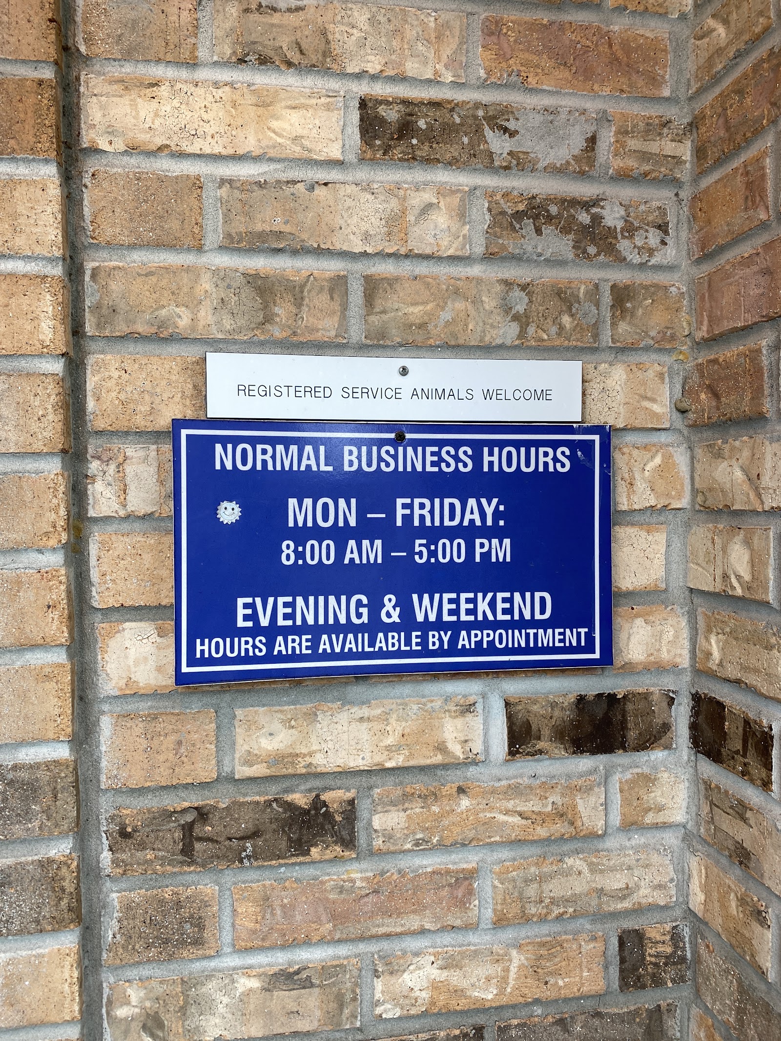 Exterior brick wall with business hours sign and service animals welcome notice mounted on building entrance