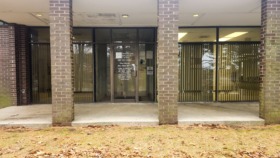 Exterior view of brick building with covered entrance, columns, and landscaped grounds with shrubs