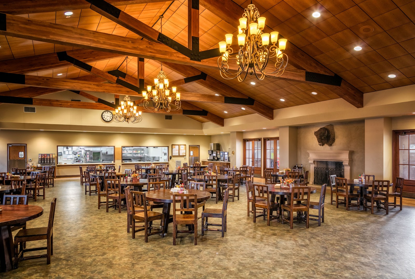 Interior dining hall with multiple round tables, wooden chairs, beamed ceiling, chandeliers, and serving area