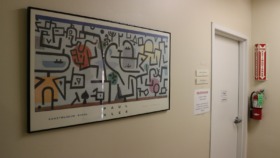 Interior corridor with metal fencing displaying student artwork including abstract designs, street scenes, and eye artwork with concrete columns