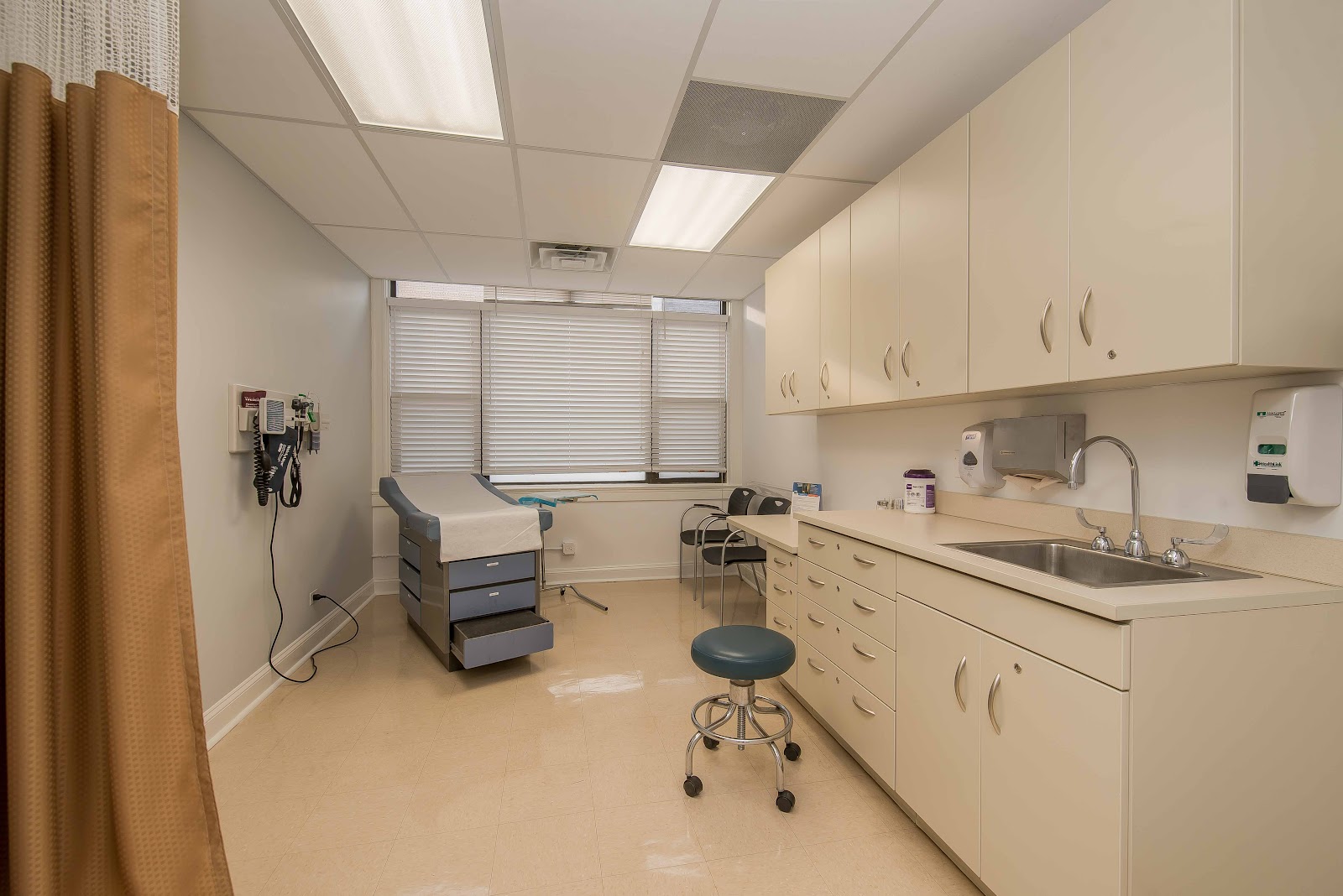 Interior clinical treatment space with examination bed, medical equipment, cabinets, sink, and window with blinds