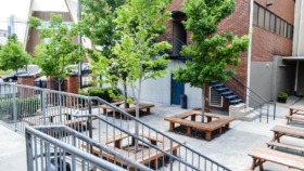 Exterior courtyard space with wooden picnic tables, metal railings, brick building facade, and mature trees providing shade