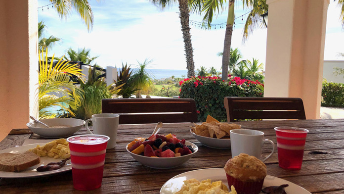 Interior dining area with wooden table, fresh fruit breakfast, tropical plants, and expansive ocean and mountain views