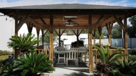 Exterior covered pavilion with wooden beams, dining tables and chairs, surrounded by tropical landscaping and palm plants