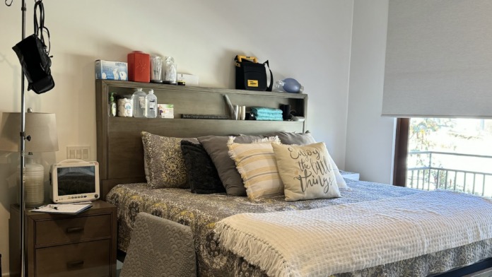Interior patient bedroom featuring bed with patterned bedding, nightstand, medical monitoring device, and window with ocean view