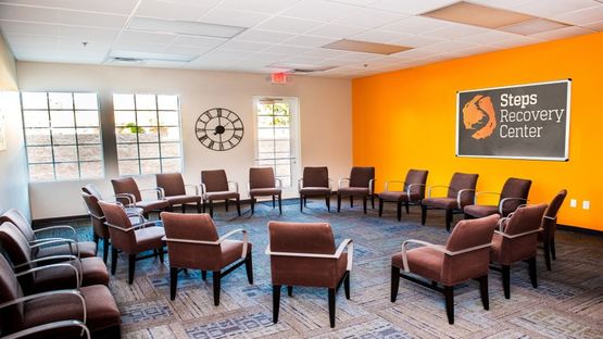 Interior community space with multiple comfortable chairs arranged in circle configuration, clock on wall, and office area visible