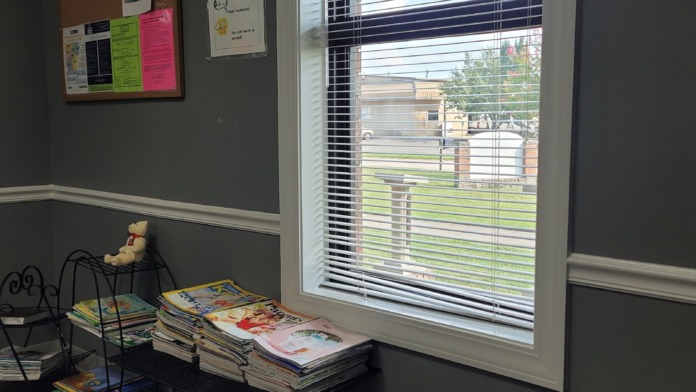 Interior room with bulletin boards, children's books and magazines on display shelves, toys, piano, and large window with blinds overlooking outdoor grounds