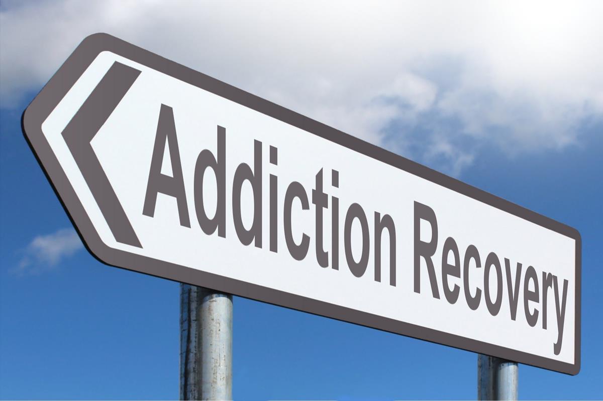 Outdoor directional sign reading 'Addiction Recovery' with left-pointing arrow against sky and clouds.
