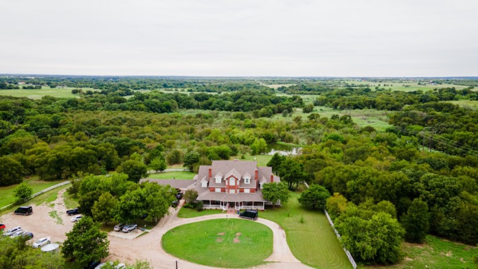 Aerial drone view of red and white farmhouse facility with parking lot, surrounding landscaping and green fields