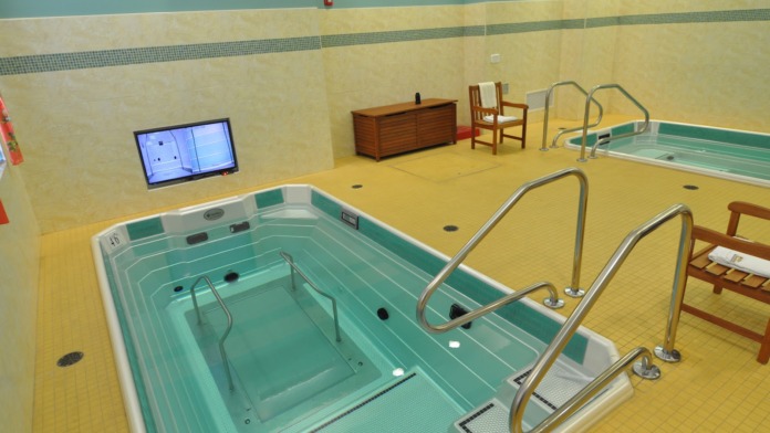 Interior therapeutic aquatic facility with two treatment pools featuring metal railings, yellow rubber flooring, and wall-mounted television monitor
