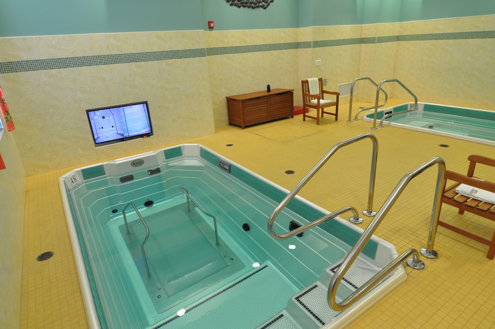 Interior therapeutic aquatic facility with two treatment pools featuring metal railings, yellow rubber flooring, and wall-mounted television monitor