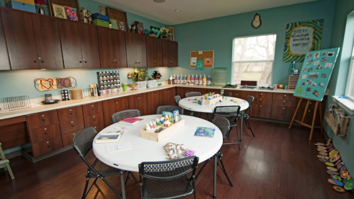 Interior creative therapy space with round tables, chairs, art supplies, storage cabinets, turquoise walls, and activity displays