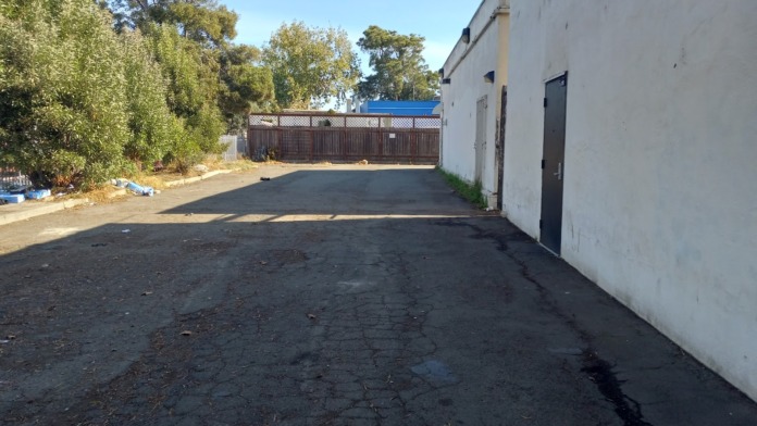 Exterior parking and grounds area with building wall, metal fencing, trees, and perimeter access