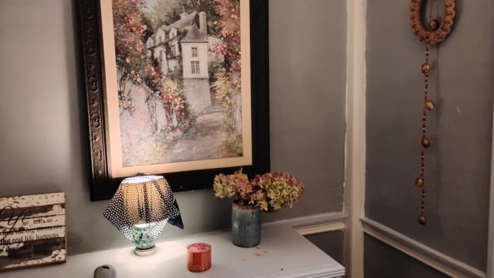 Interior bedroom featuring white nightstand with decorative lamp, framed landscape artwork, dried flowers in vase, and gray wall paneling