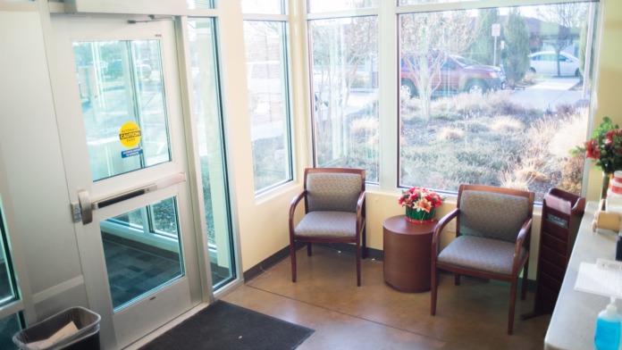 Interior lobby entry with glass doors, comfortable armchairs, decorative flowers, windows overlooking exterior parking and landscaping