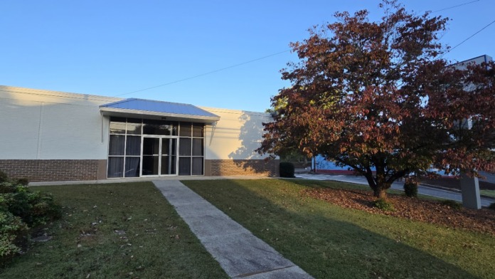 Exterior of treatment facility showing main entrance with concrete walkway, landscaped grounds, trees, and front facade of single-story white and brick building