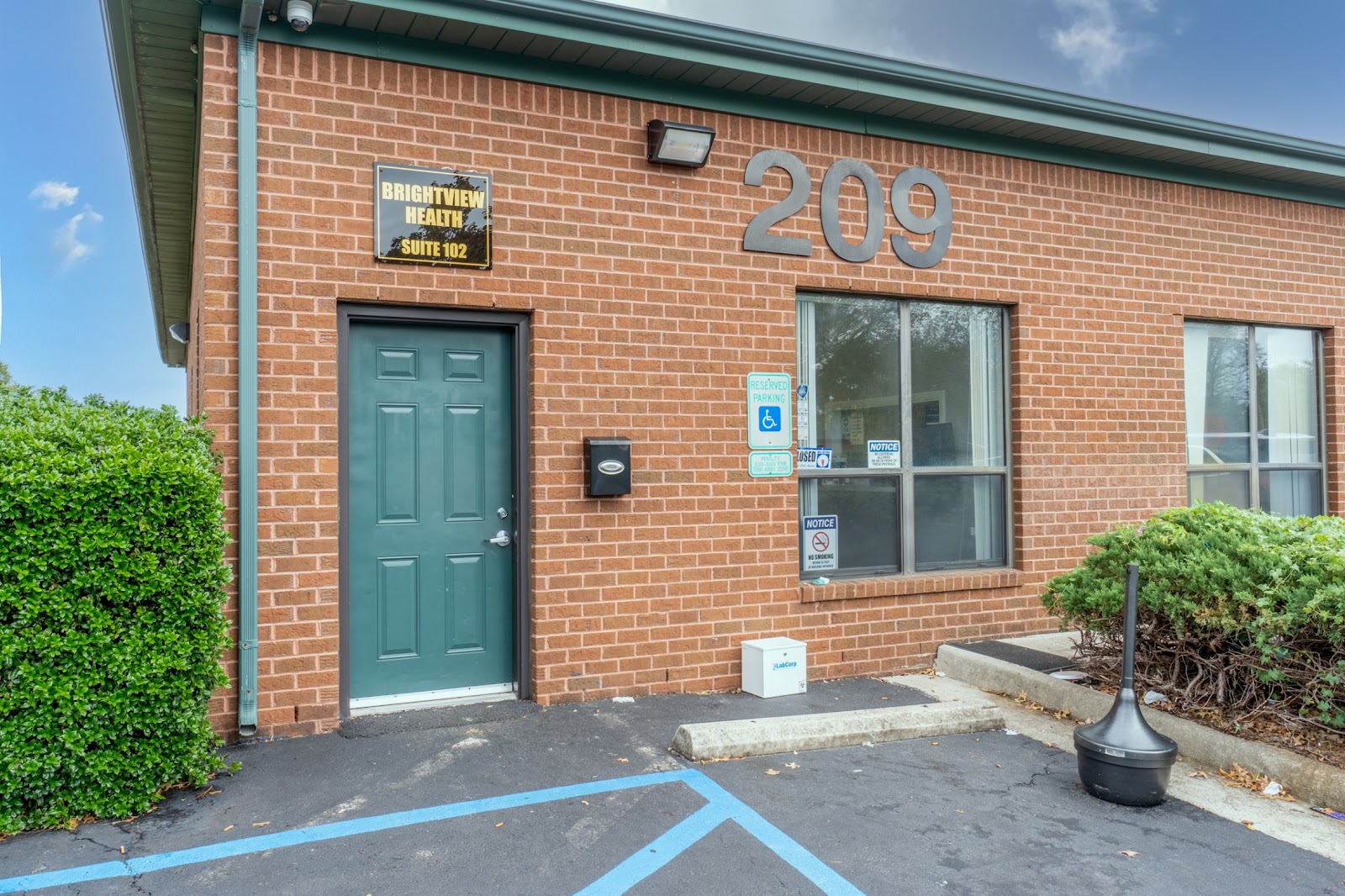 Exterior red brick building entrance showing BrightView Health Suite 102 signage, door, windows, and parking area