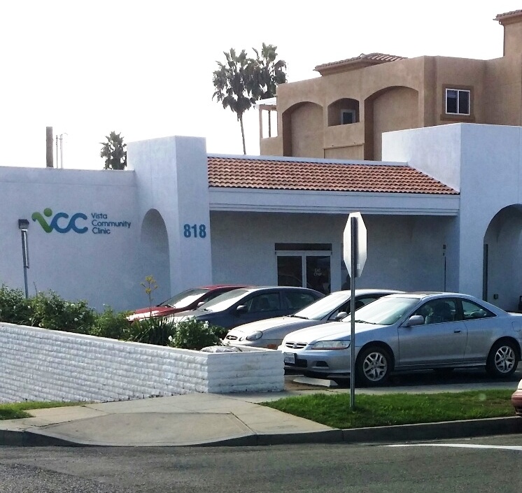 Exterior side view of white clinical building with large Vista Community Clinic signage and parking lot with multiple vehicles