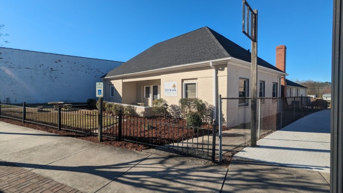 Exterior side view showing single-story beige brick building with metal railings, driveway, and adjacent structures under clear sky