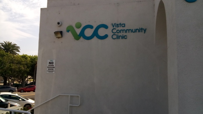 Exterior side wall of white building showing large Vista Community Clinic logo with landscaping and parking lot view