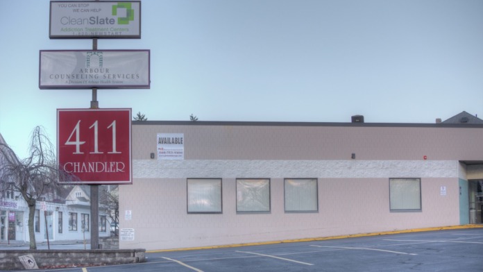 Exterior view of one-story facility building with parking spaces, address signage, and surrounding storefront area