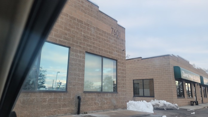 Exterior brick facade showing address number 7601, large windows with vertical blinds, and modern storefront design