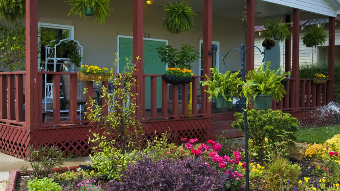 Exterior building entrance with red support columns, covered porch area, hanging plants, and flower gardens