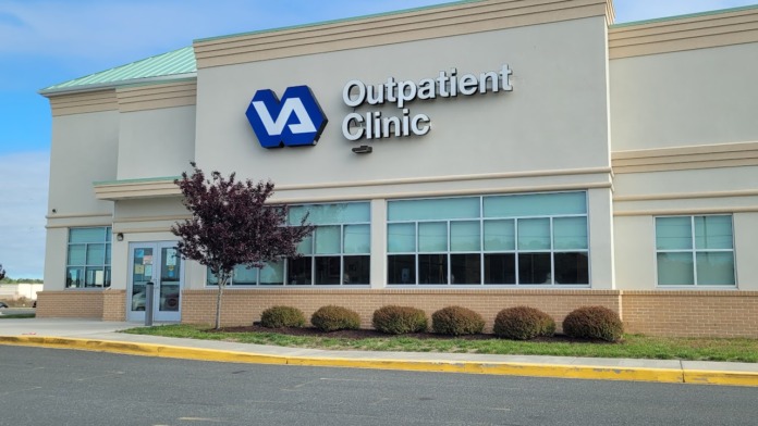 Exterior front view of VA Outpatient Clinic building with entrance doors, large windows, brick base, and landscaping shrubs
