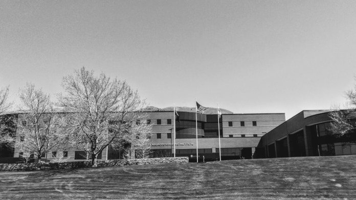 Exterior side view of facility building with evergreen landscaping and mountain backdrop in monochrome