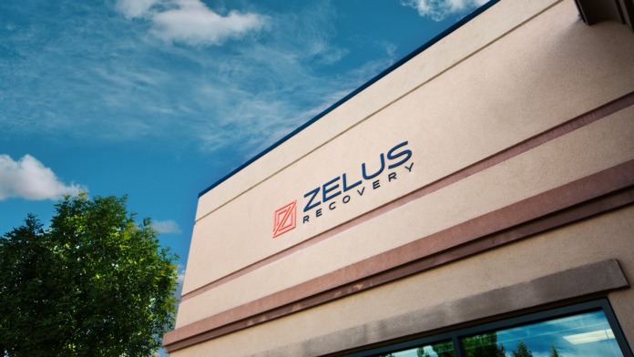 Exterior signage showing Zelus Recovery logo and text on beige building facade with dark trim against blue sky
