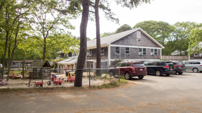 Exterior view of facility building with gray shingles, white railings, mature trees, and landscaped grounds