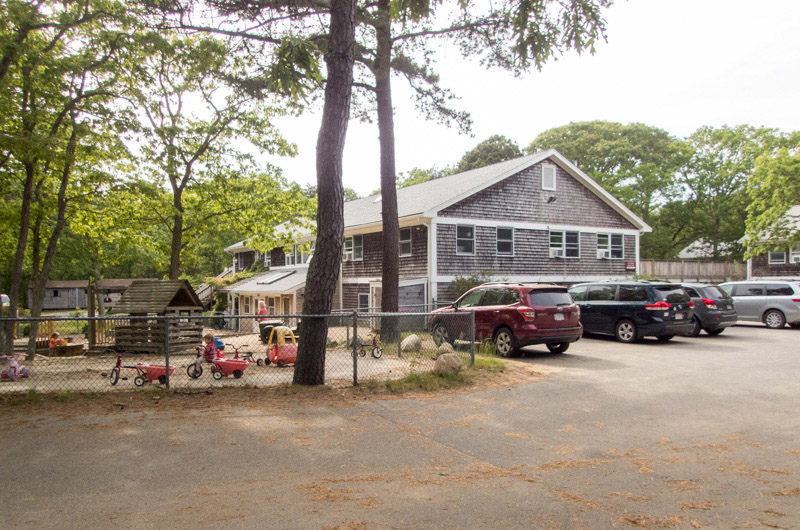 Exterior view of facility building with gray shingles, white railings, mature trees, and landscaped grounds