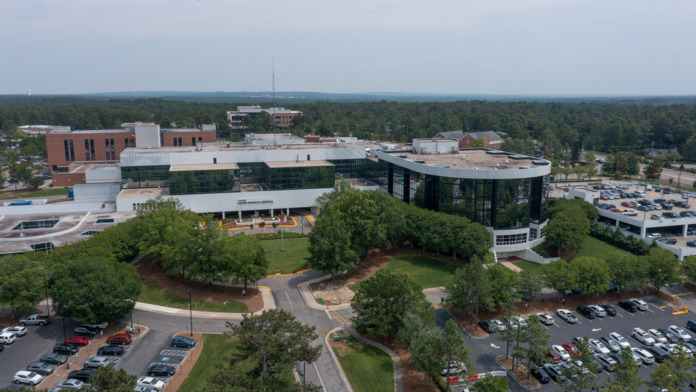 Aerial photograph of hospital campus showing multiple building structures, connected facilities, parking areas, trees, and surrounding landscape