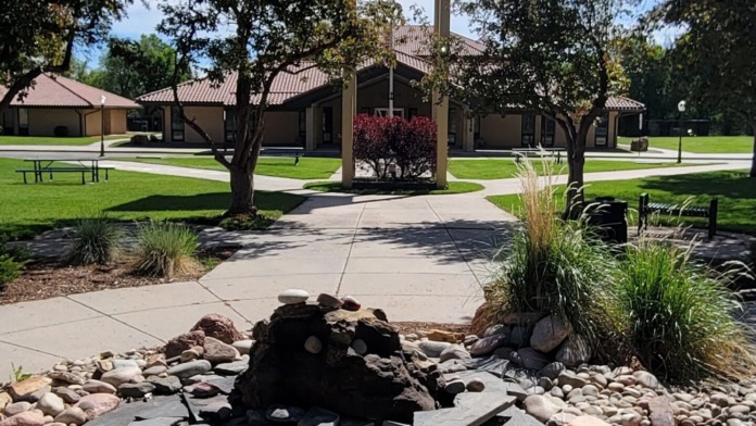 Outdoor recreational area featuring fire pit with stones, clock tower, picnic tables, and manicured grounds with mature trees