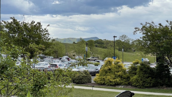 Exterior grounds view showing visitor parking lot, landscaping with flowering plants and shrubs, green lawn areas, and hospital building