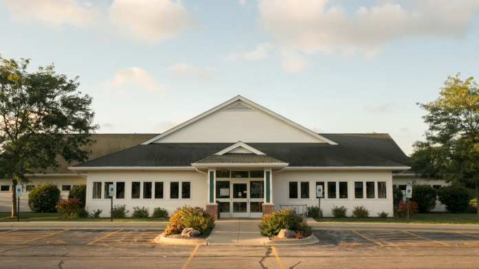 Exterior health center with white siding, landscaped grounds, center entrance portico, and asphalt parking area with yellow line markings