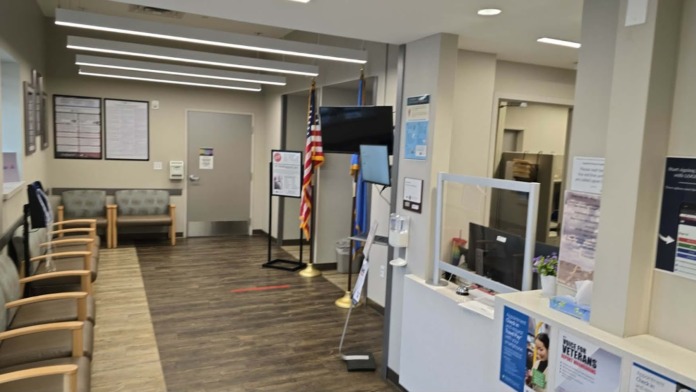 Interior reception and check-in area with white desk, computer monitors, flags, information boards, and seating along the wall