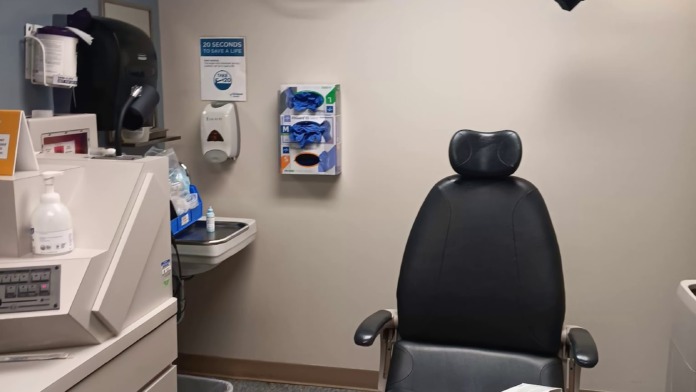 Interior medical exam room with dental/clinical chair, equipment, monitor, cabinets, and medical supplies