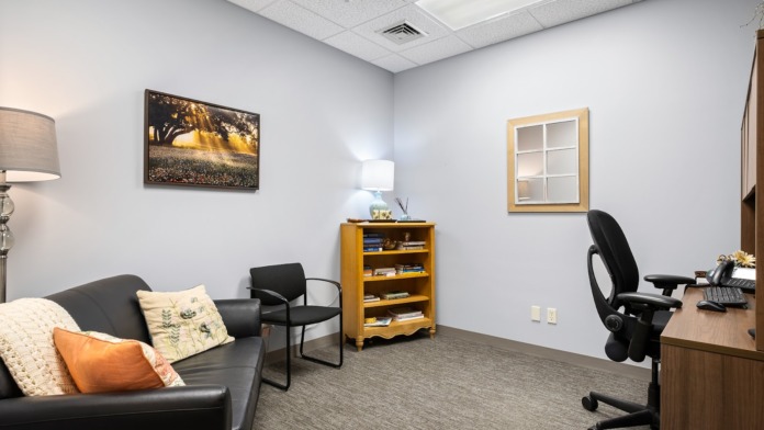 Interior office room with black sofa, wooden desk, office chair, shelving unit, and framed wall art